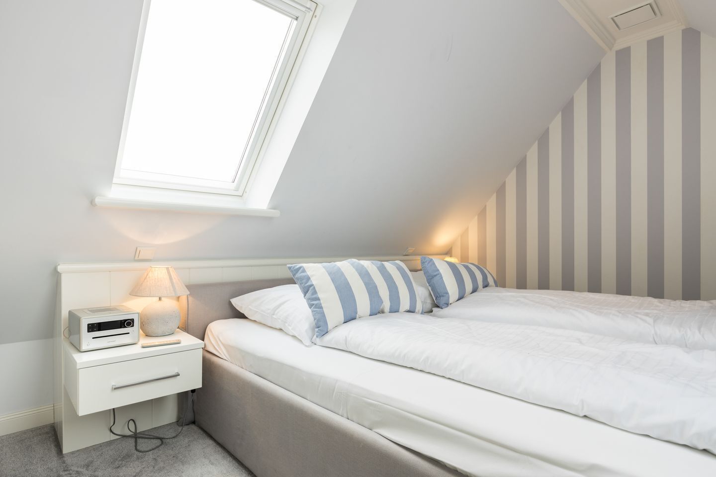 Ein Schlafzimmer mit schrägverdeckter Decke, einem Bett mit weißer Bettwäsche und gestreiften Kissen, einem Nachttisch mit Lampe und einem kleinen weißen Gerät.