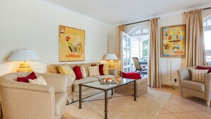 A well-lit living room with a sofa set, a coffee table, and two lamps. There are two paintings on the walls, and a window with curtains. - Nette-star, App. 14 - Einmalige Lage. Fantastischer Wellnessbereich. Viel Platz und Stil für Zwei (Sylt)