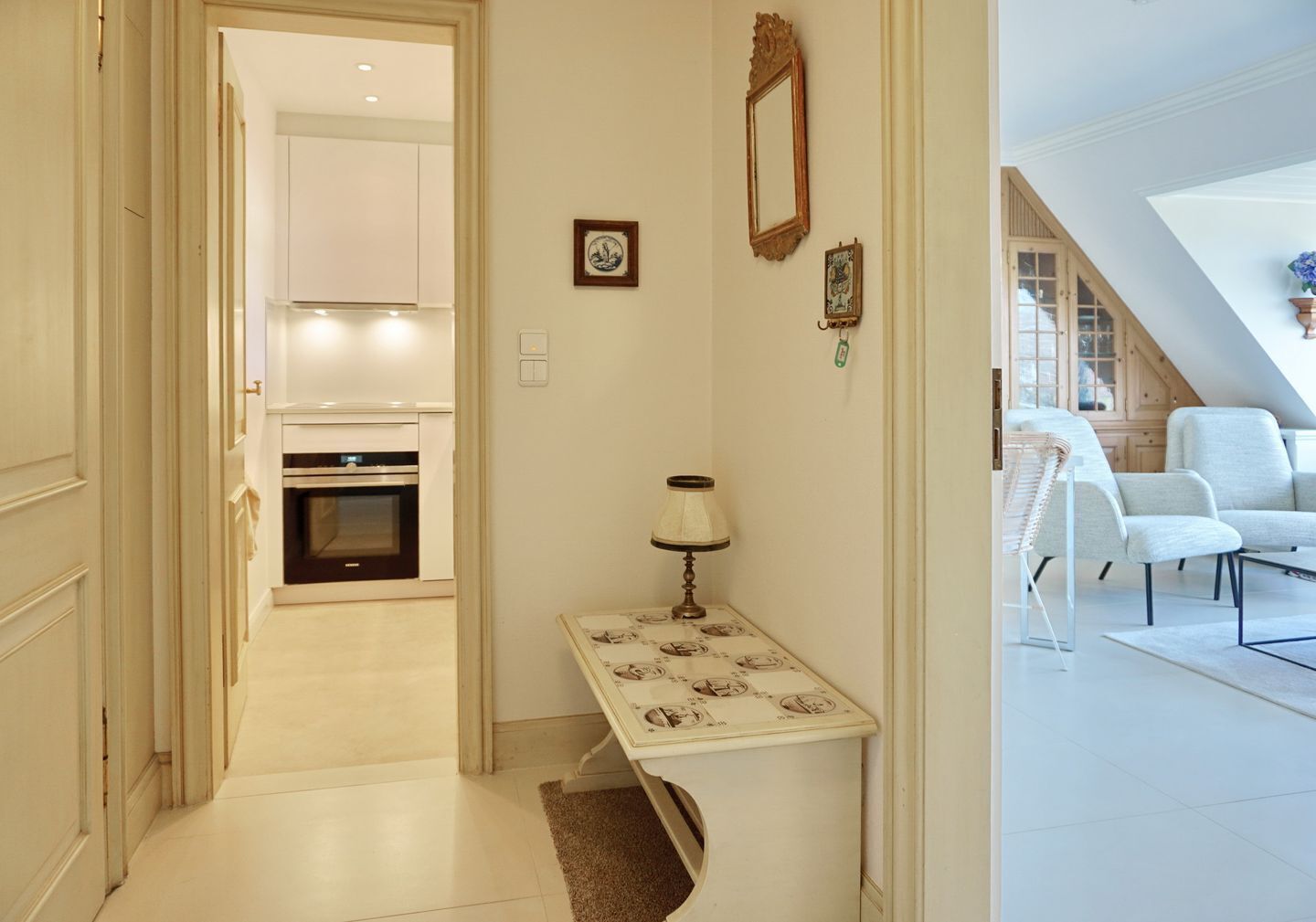 A hallway leading to a kitchen with white cabinets and a built-in oven. There is a small table with a lamp and a rug. A framed picture hangs on the wall.
