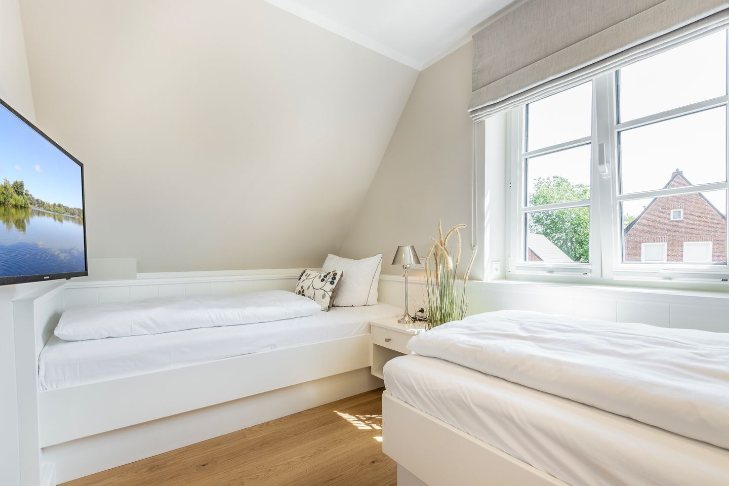 A bedroom with two beds, a TV, a window with a view of a brick building, and a bedside table with a lamp and decorative plants.