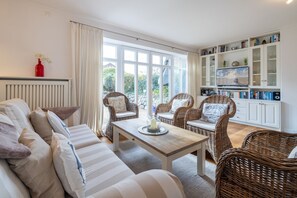 Una sala de estar acogedora con un sofá, dos sillones de mimbre, una mesa de centro de madera y una estantería llena de libros y objetos decorativos.
