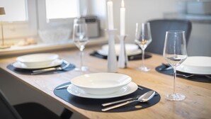 A dining table set with white plates, cutlery, and wine glasses, with candles and a modern kitchen in the background. - Mali - 3-raum Appartement im Ober- und Dachgeschoss mit ca. 75 m² Nutzfläche (Sylt)