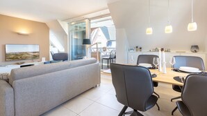 A modern living room with a grey sofa, a dining table set for four, and a TV mounted on the wall. - Mali - 3-raum Appartement im Ober- und Dachgeschoss mit ca. 75 m² Nutzfläche (Sylt)