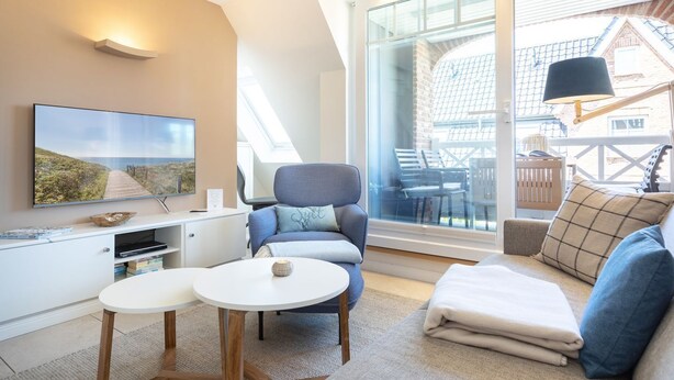 A modern living room with a large window, a flat-screen TV, a sofa, and a coffee table. - Mali - 3-raum Appartement im Ober- und Dachgeschoss mit ca. 75 m² Nutzfläche (Sylt)