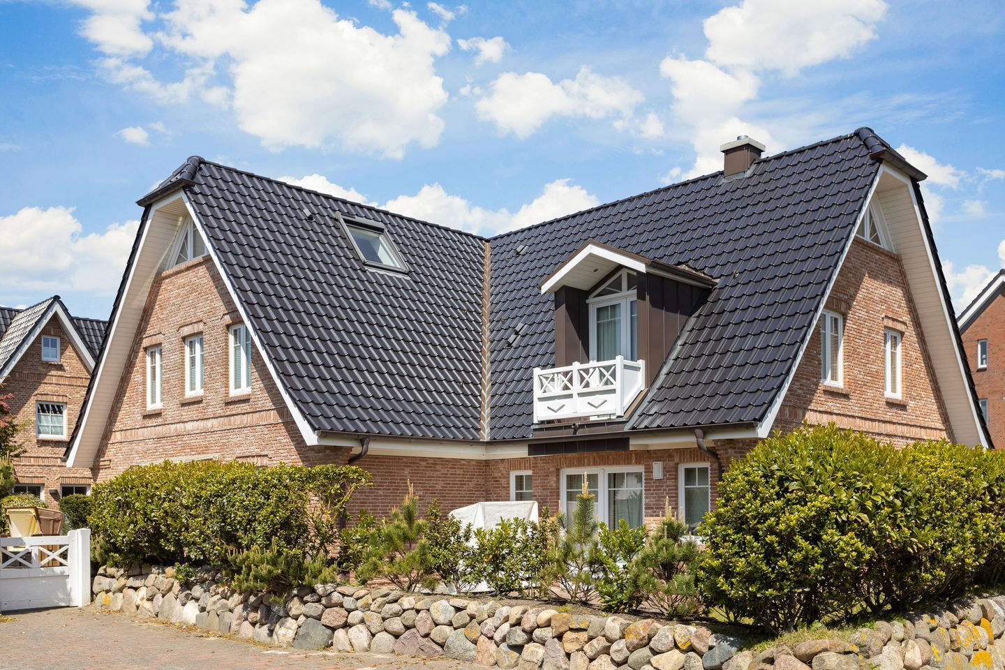 A two-story house with a steeply pitched roof, a balcony with a white railing, and a stone wall in the foreground.