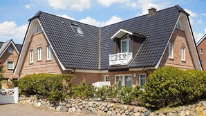 A two-story house with a steeply pitched roof, a balcony with a white railing, and a stone wall in the foreground. - Mali - 3-raum Appartement im Ober- und Dachgeschoss mit ca. 75 m² Nutzfläche (Sylt)