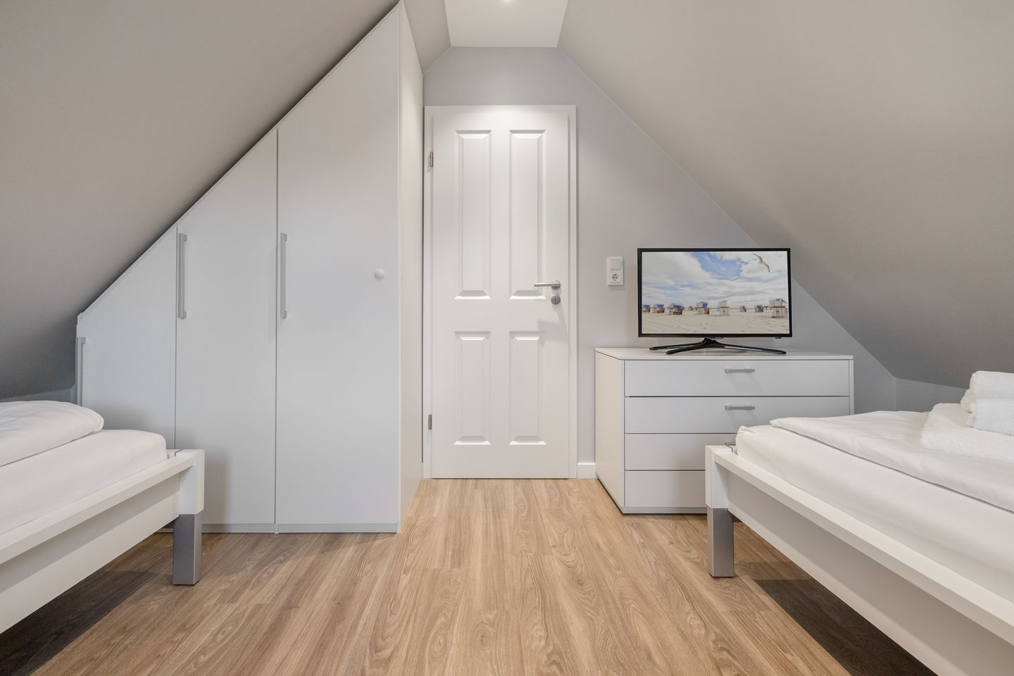 A modern bedroom with a sloped ceiling, white furniture, and a television on a dresser.