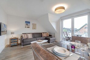 A modern living room with a grey sofa, a coffee table, and a dining table set for four. There are two paintings on the wall, a lamp on a wooden side table, and a view of buildings through large windows.