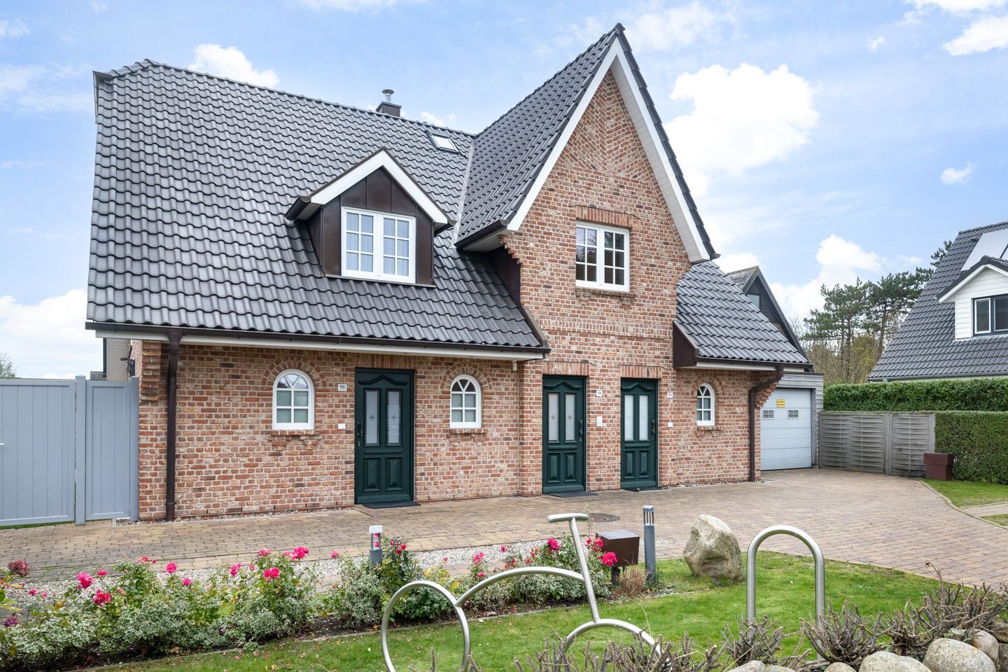 Een huis met twee verdiepingen, een gevel van baksteen, een grijs pannendak en een voortuin met bloemen en een fietsenrek van metaal.
