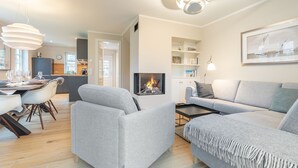 Ein modernes Wohnzimmer mit grauen Sofas, einem Kamin, einem Essbereich mit Tisch und Stühlen sowie einer Küche im Hintergrund.
