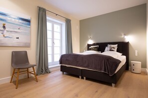Un dormitorio con cama, una silla, un cuadro y una ventana con cortinas.