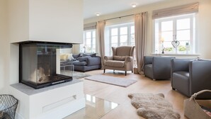 Un salon moderne avec une cheminée, des canapés gris, un fauteuil beige et de grandes fenêtres avec des rideaux.