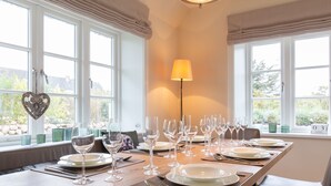 Une table à manger dressée pour six personnes, avec des verres à vin, des assiettes et des couverts, de grandes fenêtres avec des rideaux et une lampe.