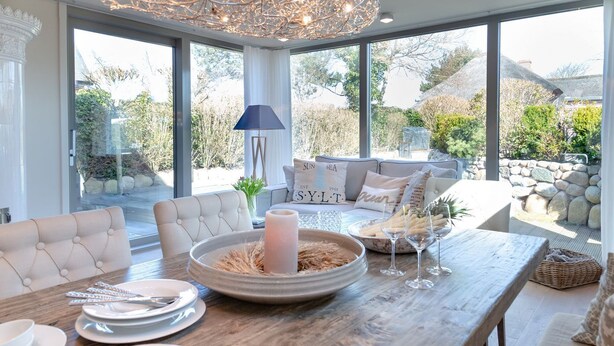 A dining area with a wooden table set for a meal, a chandelier, and a view of the garden through large windows. - Anno 1855 - Was für ein Highlight - Sowohl Außen als Auch Innen! (Sylt)