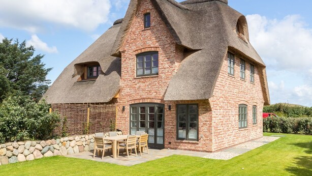 Une maison traditionnelle en briques, avec un toit de chaume, un mur en pierre et une terrasse agrémentée d’une table et de chaises.