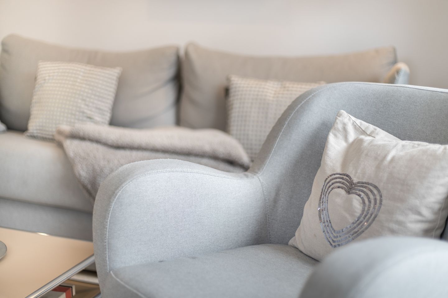 A grey armchair with a heart-shaped pillow and a grey sofa with patterned cushions in a living room.
