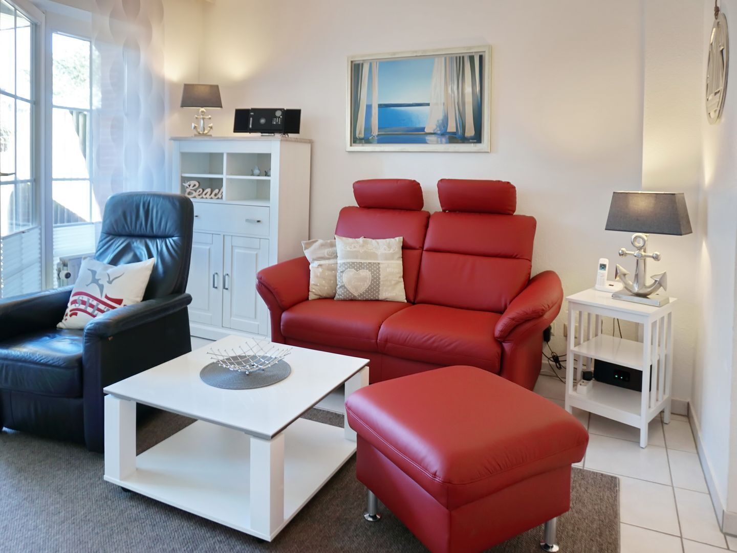 Un salon avec un fauteuil en cuir noir, un canapé rouge, une table basse blanche et une petite table d’appoint blanche avec une lampe.