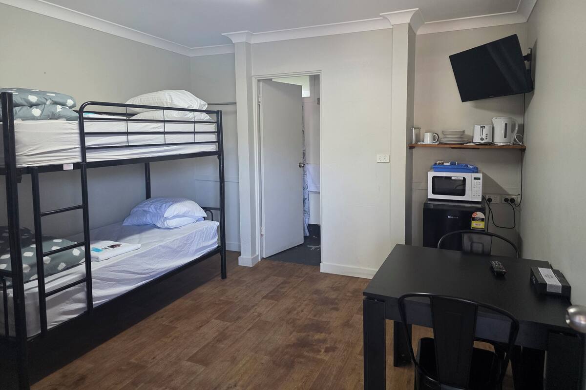 Une chambre de dortoir équipée de lits superposés, d’un bureau, d’une chaise, d’un four à micro-ondes et d’un téléviseur à écran plat fixé au mur.