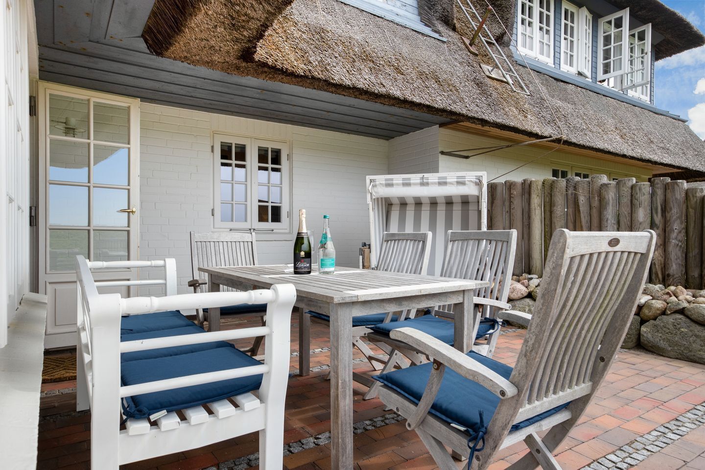 Une terrasse extérieure avec une table et des chaises, une bouteille de champagne et une structure dotée d’un toit de chaume.
