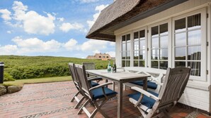 Une terrasse avec une table et des chaises, un toit de chaume et une vue sur une colline et un bâtiment au loin.