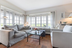Ett rymligt vardagsrum med en grå soffa, ett soffbord i glas och en vit fåtölj. Stora fönster med gardiner släpper in naturligt ljus och ger utsikt över omgivningen.