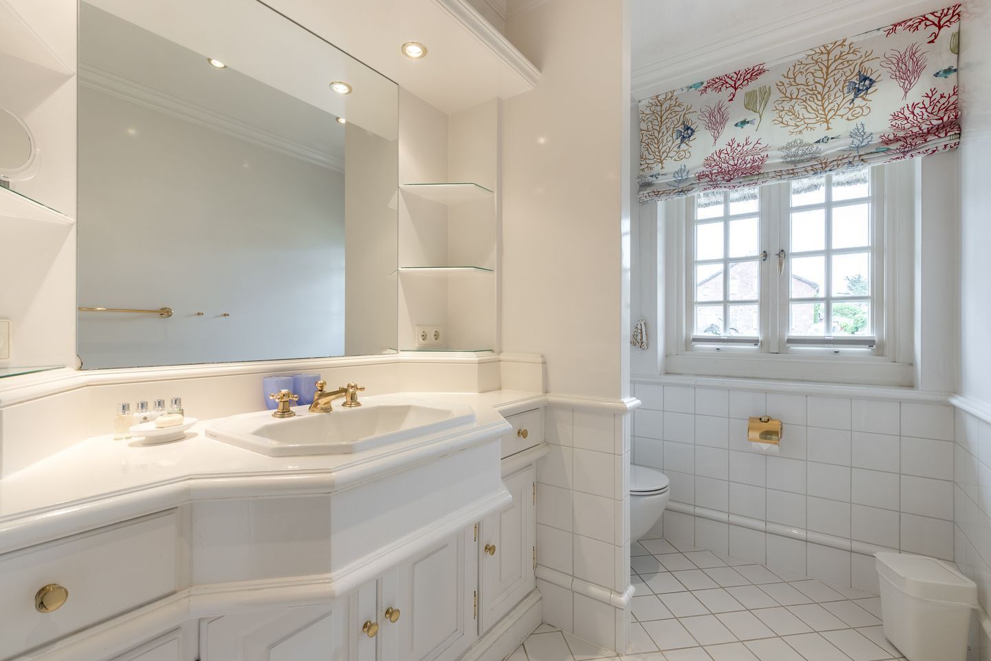 A bathroom with a large mirror, a sink with gold fixtures, and a window with a floral curtain.