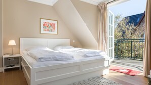 Ein Schlafzimmer mit einem weißen Bett, einem Nachttisch mit Lampe, einem gerahmten Bild an der Wand und einem Balkon mit rotem Teppich.