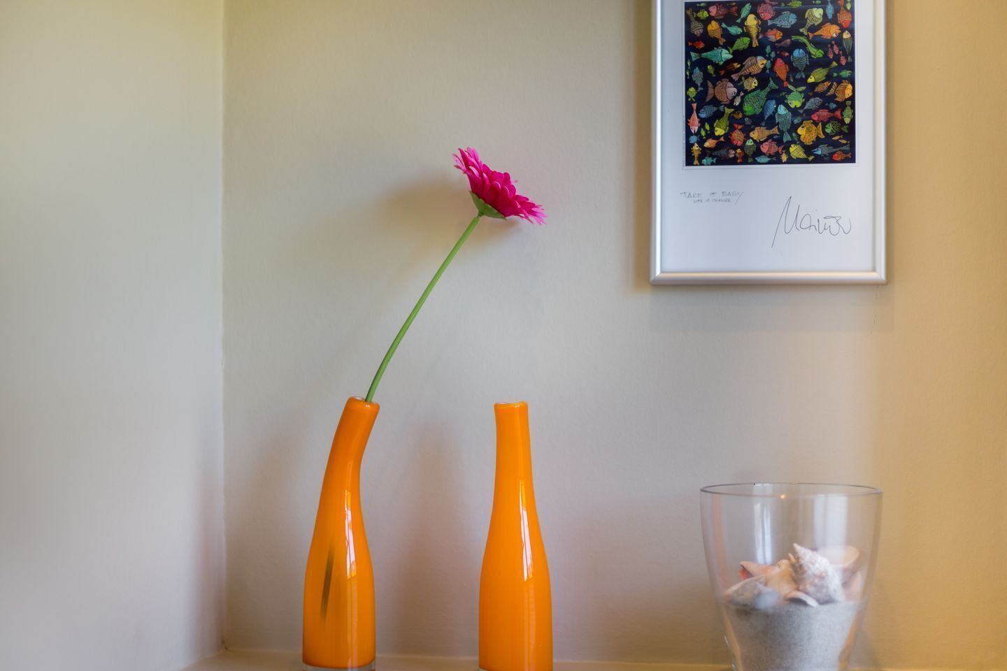 Dos jarrones naranjas con una flor rosa sobre una estantería, una obra de arte enmarcada y un recipiente de vidrio con objetos.