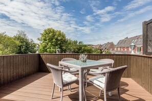 Una terraza de madera con mobiliario de mimbre y una mesa de vidrio, rodeada por una valla de madera y árboles.