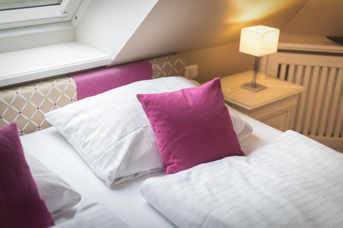A neatly made bed with white and pink pillows, a bedside lamp, and a window with a view of the outside.