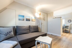 Un salon moderne avec un canapé gris, une petite table blanche et une lampe. Trois tableaux encadrés ornent le mur, et une porte est décorée d’un ornement suspendu.