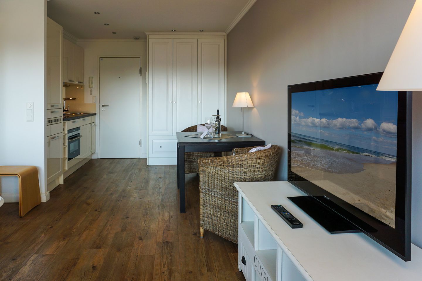 Eine moderne Küche mit weißen Schränken, ein Essbereich mit Korbmöbeln und ein großer Flachbildfernseher, der eine Strandansicht zeigt.