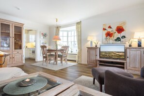 A spacious living room with a wooden dining table, comfortable seating, a television, and a large window providing natural light. - Christa, App. 1 - First-class Suiten in Bestlage (Wenningstedt-Braderup (Sylt))