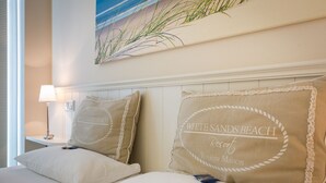 A hotel room with a bed, two pillows with 'WHITE SANDS BEACH Resort' text, a nightstand with a lamp, and a painting of beach grasses. - Landhaus Berthin-bleeg, App. 5 Strandleben - Top Lage. Beach-style Ausstattung. Viel Platz. Alles (Wenningstedt-Braderup (Sylt))