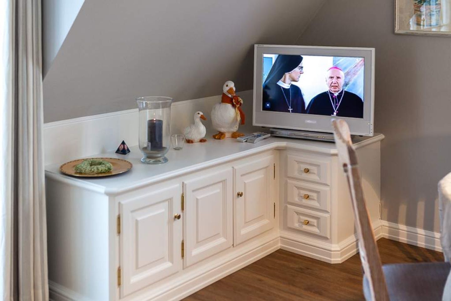Habitación de esquina con una televisión sobre un mueble blanco, un candelabro de vidrio y objetos decorativos.