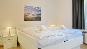 A hotel room with a white bed, two bedside tables with lamps, a framed picture on the wall, and a window with curtains. - Simokat, Eg-rechts - Sylter Süden. Verbringen Sie Schöne Ferien in Wenningstedt (Wenningstedt-Braderup (Sylt))