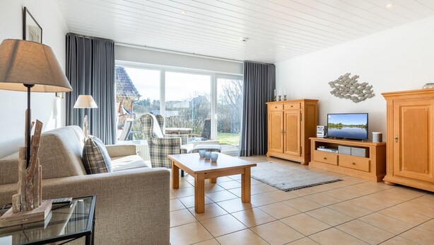 A spacious living room with a sofa, armchair, coffee table, and TV stand. Large windows offer a view of the outdoors. - Simokat, Eg-rechts - Sylter Süden. Verbringen Sie Schöne Ferien in Wenningstedt (Wenningstedt-Braderup (Sylt))