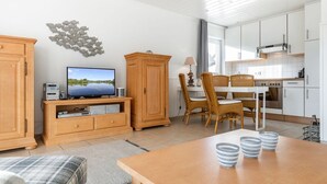 A modern living room with a TV, wooden cabinets, a dining area with wicker chairs, and a kitchen with white cabinets. - Simokat, Eg-rechts - Sylter Süden. Verbringen Sie Schöne Ferien in Wenningstedt (Wenningstedt-Braderup (Sylt))