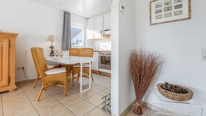 A modern kitchen with white cabinets, a white countertop, and a stainless steel oven. There is a dining area with a white table and wicker chairs. - Simokat, Eg-rechts - Sylter Süden. Verbringen Sie Schöne Ferien in Wenningstedt (Wenningstedt-Braderup (Sylt))
