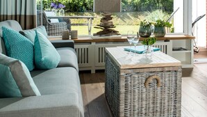 A modern living room with a grey sofa, a wicker coffee table, and a view of the garden through large windows.