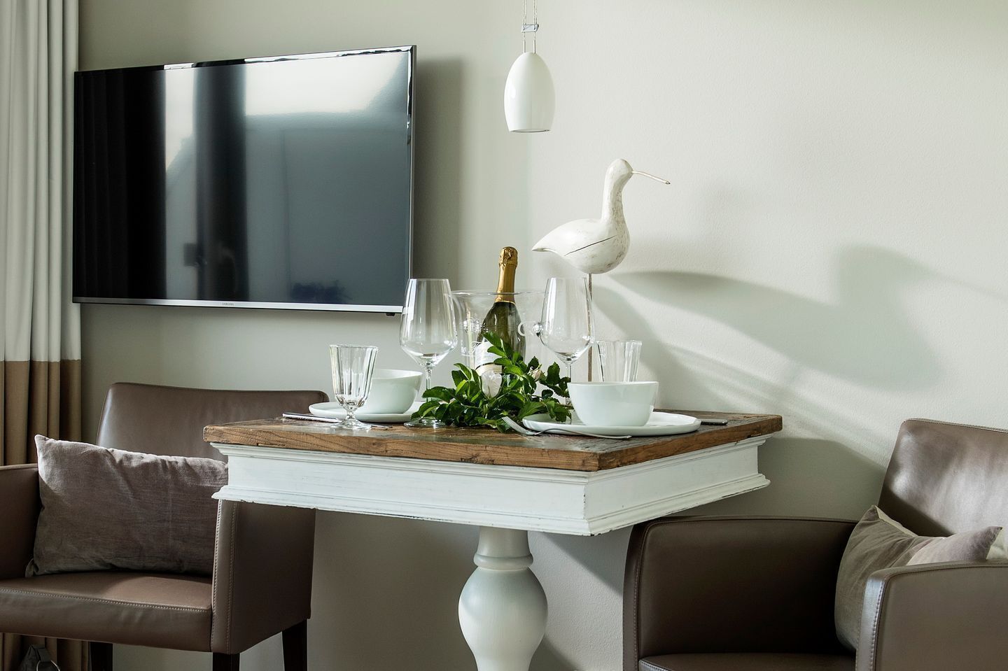 Une table à manger dotée d’un pied central blanc, entourée de chaises en cuir marron. Un téléviseur à écran plat est fixé au mur, au-dessus de la table.