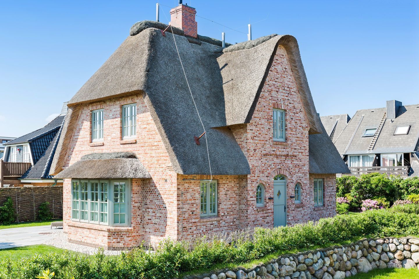 A traditional brick house with a steeply pitched thatched roof, multiple windows, and a stone wall in the foreground.
