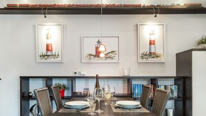 A dining area with a wooden table set for four, surrounded by wicker chairs. Above the table, there are three framed artworks depicting lighthouses. A bookshelf with books and decorative items is to the right. - Landhaus Berthin-bleeg, App. 8 Di Füürtoorn - Top Lage. Design-ausstattung. Alles auf Einer Ebene (Wenningstedt-Braderup (Sylt))