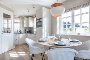 A modern kitchen with white cabinetry, a dining area with round tables and white chairs, and large windows providing natural light. - Lille Wung - Ruhige und Dennoch Zentrale Lage? (Wenningstedt-Braderup (Sylt))