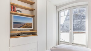 Un dormitorio con cama, televisor montado en la pared, ventana con vista a un paisaje nevado y estantería con libros.