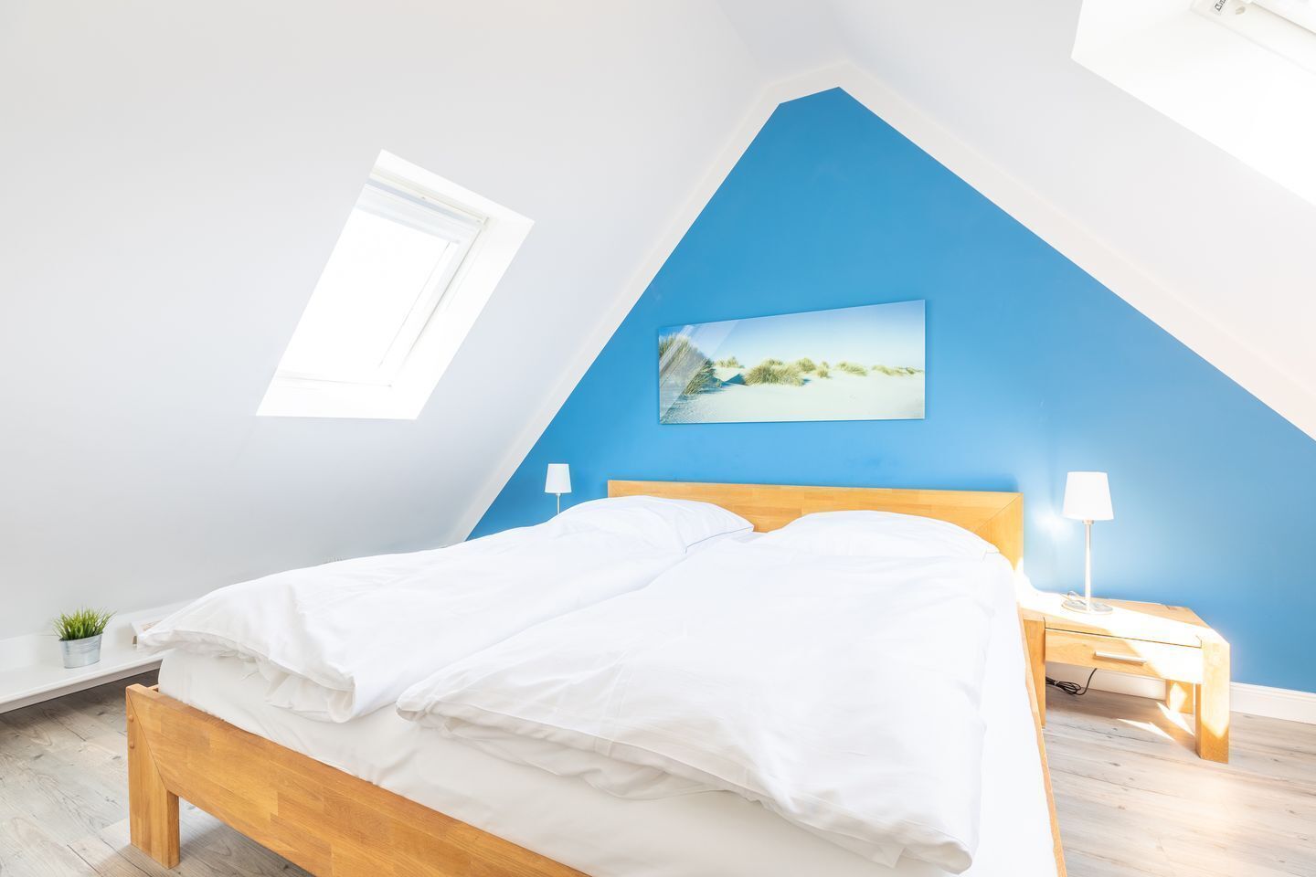 A bedroom with a sloped ceiling, a bed with white bedding, a nightstand with a lamp, and a painting of a beach scene.
