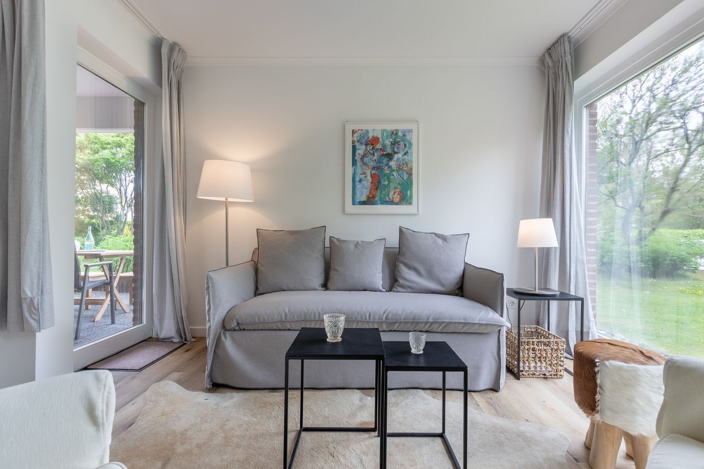 Uma sala de estar moderna com um sofá cinzento, duas mesas pretas, uma luminária e um quadro na parede.