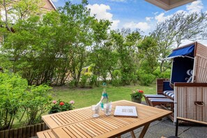 Une table en bois avec une bouteille et des verres, une chaise en osier et un espace salon abrité, le tout dans un cadre de jardin.