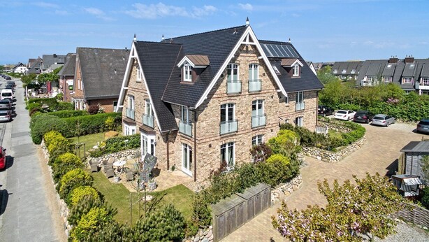 A large stone house with a steeply pitched roof, multiple balconies, and a driveway with parked cars. - Landhaus Berthin-bleeg, App. 7 Lichtblick OG Rechts - Zwei Raum Appartement im 1. OG auf Einer Ebene (Wenningstedt-Braderup (Sylt))
