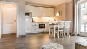 A modern kitchen with white cabinets, a dining area with a table and chairs, and a large window with curtains. - Landhaus Berthin-bleeg, App. 7 Lichtblick OG Rechts - Zwei Raum Appartement im 1. OG auf Einer Ebene (Wenningstedt-Braderup (Sylt))